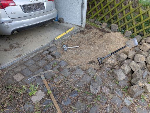 Werkzeuge und Sand liegen auf einem gepflasterten Weg neben einem Auto in einer Garage.